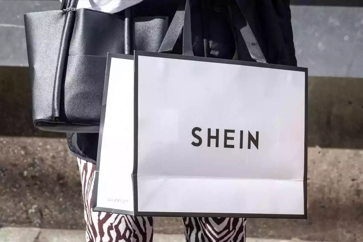 Foto de una mujer de la que solo se ven las piernas y que lleva un bolso negro y una bolsa blanca grande en la que pone Foto de una mujer de la que solo se ven las piernas y que lleva un bolso negro y una bolsa blanca grande en la que pone
