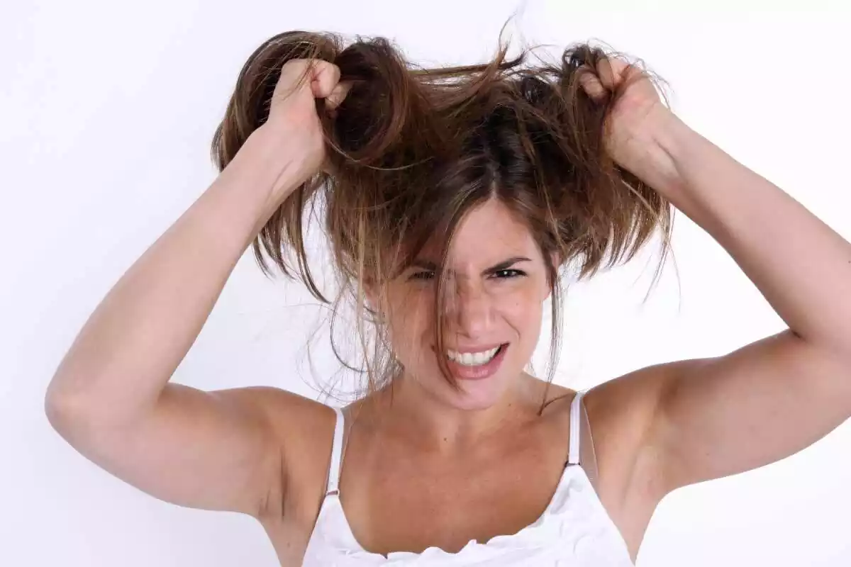 Foto de una mujer con camiseta blanca, se sujeta el pelo con las manos y tiene cara de desesperación por no poder peinarse bien Foto de una mujer con camiseta blanca, se sujeta el pelo con las manos y tiene cara de desesperación por no poder peinarse bien
